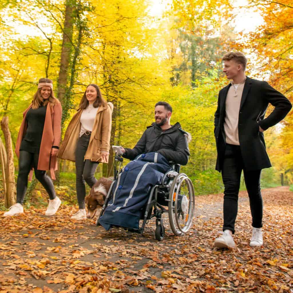 Vier junge Menschen gehen durch den Herbstwald. Einer von ihnen sitz im Rollstuhl und hat die Rolling Buttons Rollstuhldecke an. Sie unterhalten sich und lachen.