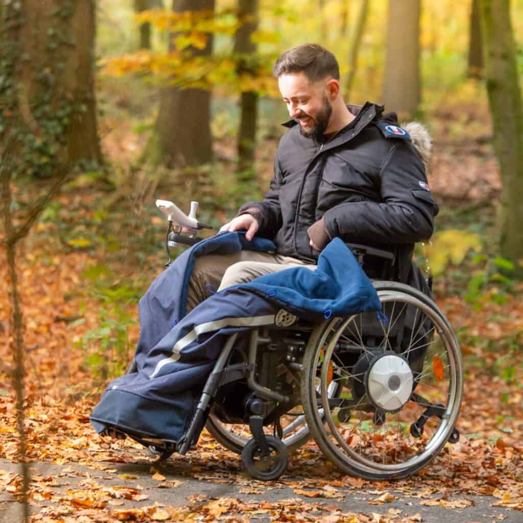 Mann im Rollstuhl schließt die Rolling Buttons Rollstuhldecke über seinen Beinen. Er befindet sich im Herbstwald.