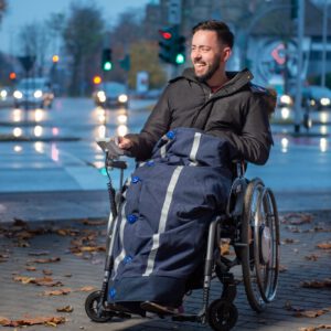 Junger Mann im Rollstuhl and befahrener Straße in der Dämmerung. Rollstuhldecke schützend über den Beinen